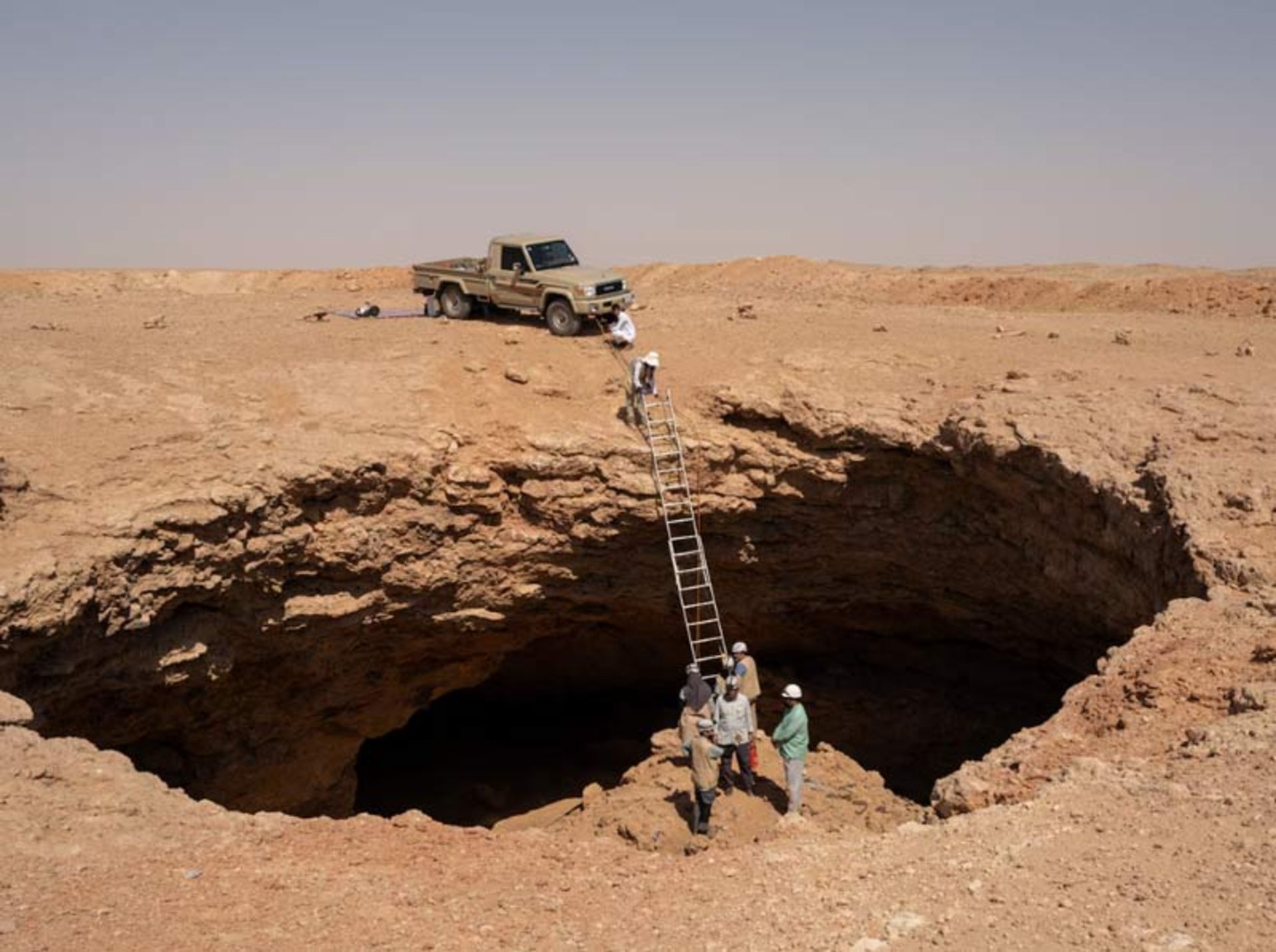 A car at the edge of a desert pit and a ladder leading io its dark depths